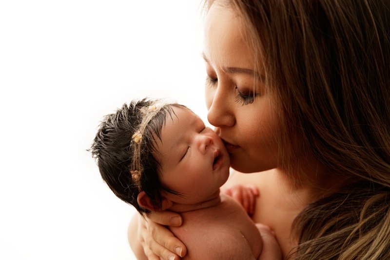 Mother kissing her newborn daughter's cheek, backlit newborn portrait
