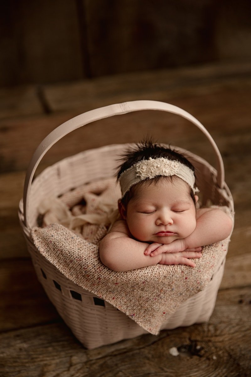 Newborn girl with a lace headband asleep in a basket, chin resting on her hands