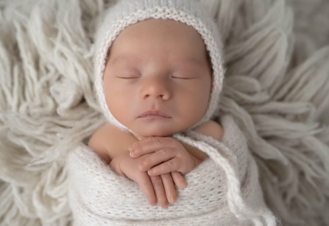 Newborn in a cream knit bonnet with hands folded, wrapped in a soft blanket
